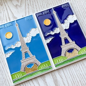 Op de afbeelding: Twee blauwe en witte keramische tegels met de Eiffeltoren en de woorden "Tour Eiffel" en "Paris".