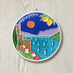 Può includere: Una targa rotonda in ceramica con sfondo blu che raffigura una scena lacustre con barche a vela, un castello e un albero di limone. Il testo "Lago di Garda" è scritto in bianco sulla targa.