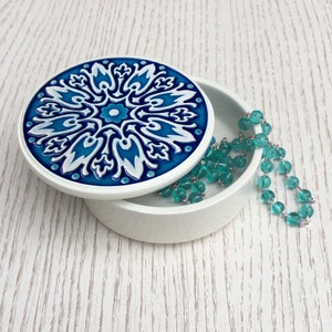 May include: A white ceramic trinket box with a blue and white patterned lid. The lid features a floral design with intricate details. A turquoise beaded necklace is resting on the box.