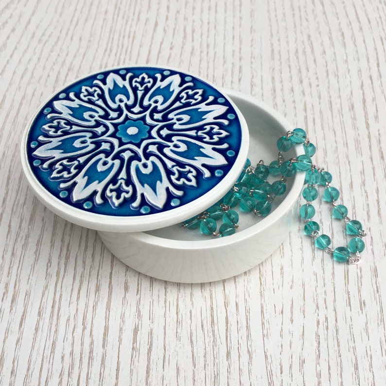 May include: A white ceramic trinket box with a blue and white patterned lid. The lid features a floral design with intricate details. A turquoise beaded necklace is resting on the box.
