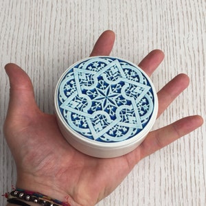 May include: A white ceramic trinket box with a blue and white geometric design. The design features a starburst pattern with intricate details.