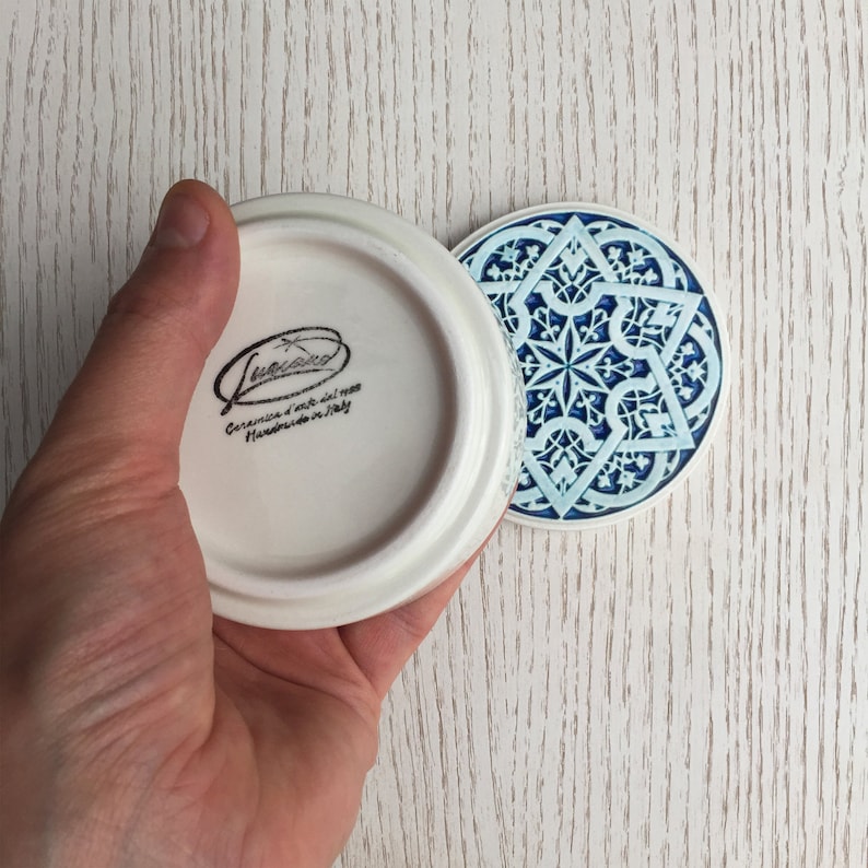 May include: A white ceramic lid with a blue and white geometric design on the bottom. The lid has the text "Ceramica d'arte del mese Handmade in Italy" on the bottom.