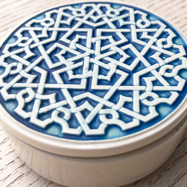 May include: A white ceramic trinket box with a blue and white geometric design on the lid. The design is intricate and features a repeating pattern of interlocking shapes.