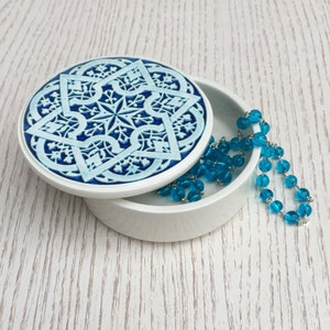 May include: A white ceramic trinket box with a blue and white geometric design on the lid. The box is open and a turquoise blue glass necklace is inside.