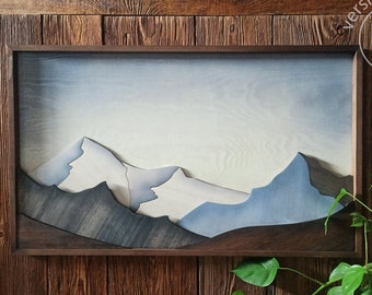 Puede incluir: Una pieza de arte de pared de madera que representa una cordillera con un esquema de color azul y blanco. El arte está enmarcado en un marco de madera oscura y está montado en una pared de madera.
