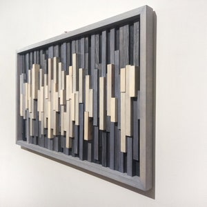 May include: A rectangular wooden wall art piece with a gray frame. The art is made of various sized wooden slats in shades of gray and beige, arranged in a vertical pattern.