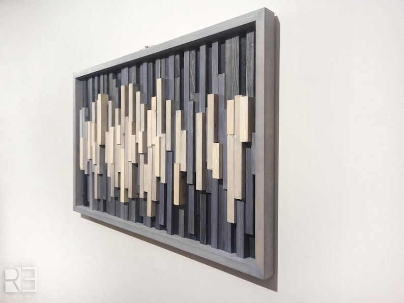 May include: A rectangular wooden wall art piece with a gray frame. The art is made of various sized wooden slats in shades of gray and beige, arranged in a vertical pattern.