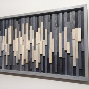 May include: A gray wood wall art piece with a wave-like pattern made of various shades of gray and beige wood pieces. The art is framed in a gray wood frame.