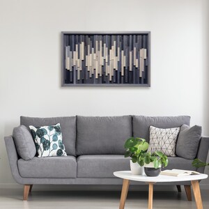 May include: A gray sofa with two pillows, one with a green leaf pattern and the other with a geometric pattern, sits in front of a wall with a wooden wall art piece featuring a geometric pattern of light and dark gray wood.