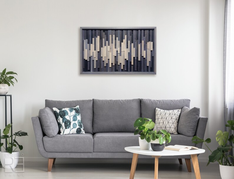 May include: A gray sofa with two pillows, one with a green leaf pattern and the other with a geometric pattern, sits in front of a wall with a wooden wall art piece featuring a geometric pattern of light and dark gray wood.