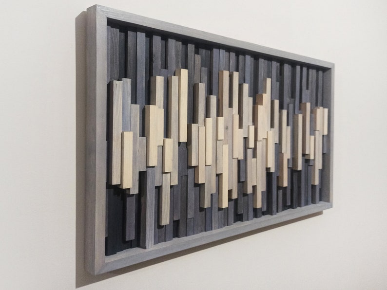 May include: A gray wood wall art piece with a geometric design. The piece is made up of various sized wood strips in shades of gray, beige, and brown. The wood strips are arranged in a wave-like pattern.