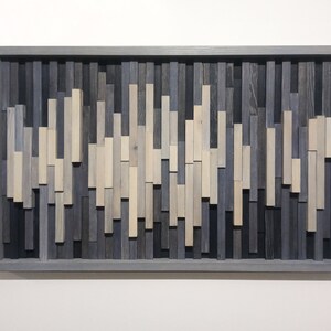 May include: A framed wall art piece made of wood with a wave-like pattern. The wood is stained in shades of gray and white.