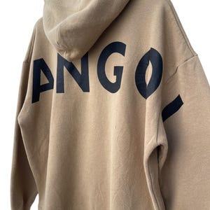 May include: A tan hoodie with a hood and long sleeves. The word "ANGO" is printed in large, black letters across the back of the hoodie. The hoodie is made of a soft, comfortable material.