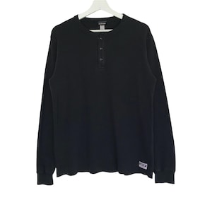 Puede incluir: Camisa Henley negra de manga larga con tapeta de tres botones. La camisa está hecha de una tela texturizada y tiene una pequeña etiqueta en la parte inferior izquierda. La camisa está colgada en una percha blanca.