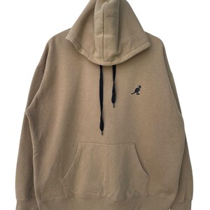 May include: A tan hoodie with a kangaroo logo on the chest. The hoodie has a drawstring, a front pocket, and a hood. The kangaroo logo is black. The hoodie is made of a soft material.