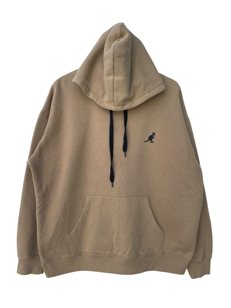 May include: A tan hoodie with a kangaroo logo on the chest. The hoodie has a drawstring, a front pocket, and a hood. The kangaroo logo is black. The hoodie is made of a soft material.