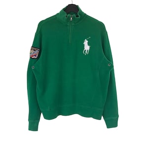 May include: Green sweatshirt with a quarter-zip collar and a white polo player logo on the chest. Features a patch on the left sleeve and ribbed cuffs. The sweatshirt is hanging on a black hanger.