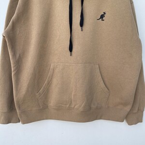 May include: A tan hoodie with a kangaroo logo on the chest. The hoodie has a front pocket, a hood, and black drawstrings. The kangaroo logo is black. The hoodie is made of a soft material.