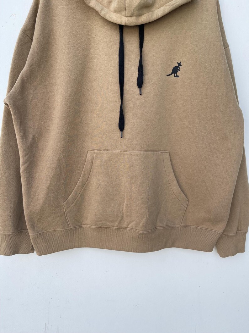 May include: A tan hoodie with a kangaroo logo on the chest. The hoodie has a front pocket, a hood, and black drawstrings. The kangaroo logo is black. The hoodie is made of a soft material.