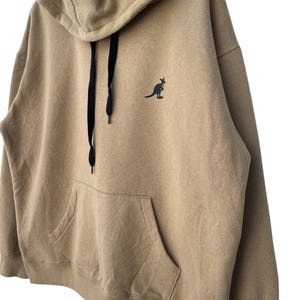 May include: A tan hoodie with a kangaroo logo on the chest. The hoodie has a drawstring hood, a large front pocket, and black drawstrings. The kangaroo logo is black. The hoodie is made of a soft, comfortable material.