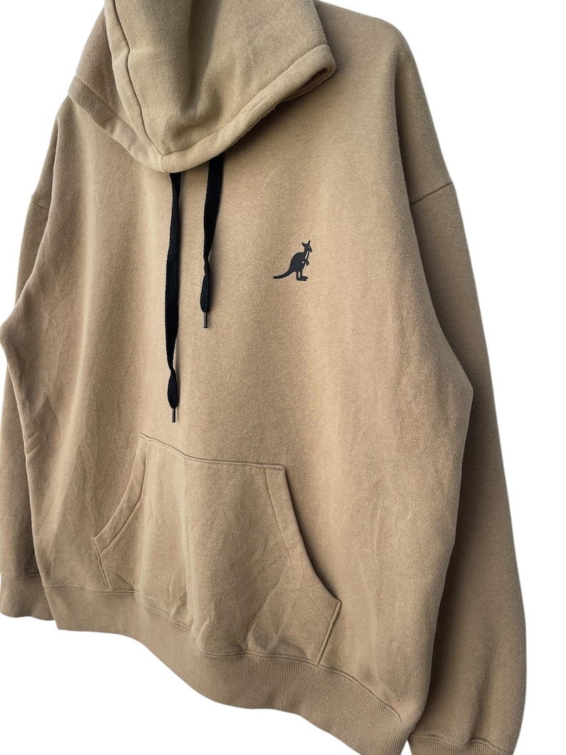 May include: A tan hoodie with a kangaroo logo on the chest. The hoodie has a drawstring hood, a large front pocket, and black drawstrings. The kangaroo logo is black. The hoodie is made of a soft, comfortable material.