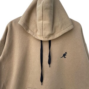 May include: A tan hoodie with a kangaroo logo on the chest. The hoodie features a large hood with black drawstrings and a front pocket. The material appears to be a soft, comfortable fabric, suitable for casual wear.