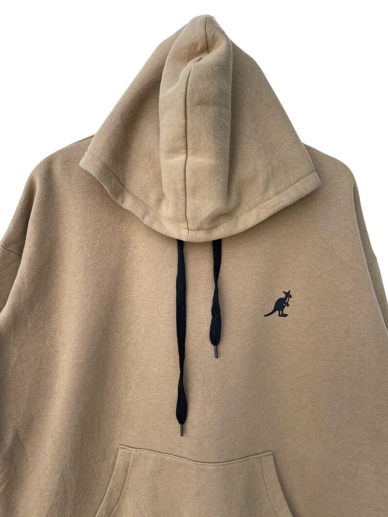 May include: A tan hoodie with a kangaroo logo on the chest. The hoodie features a large hood with black drawstrings and a front pocket. The material appears to be a soft, comfortable fabric, suitable for casual wear.