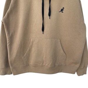 May include: A tan hoodie with a kangaroo logo on the chest. The hoodie has a drawstring hood, a kangaroo pocket, and black drawstrings. The kangaroo logo is black. The hoodie is made of a soft, comfortable material.