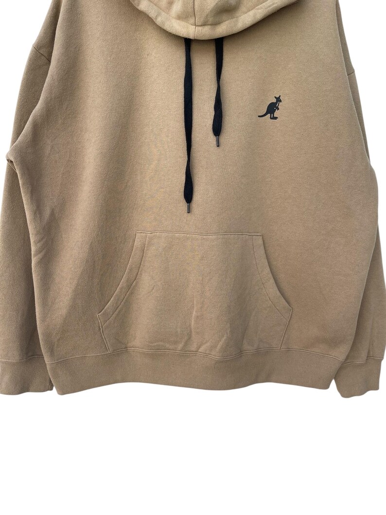 May include: A tan hoodie with a kangaroo logo on the chest. The hoodie has a drawstring hood, a kangaroo pocket, and black drawstrings. The kangaroo logo is black. The hoodie is made of a soft, comfortable material.