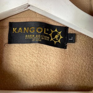 May include: Close-up of a tan-colored hoodie with a black label. The label reads "KANGOL" in gold lettering, with "BORN BRITISH ESTABLISHED IN 1938" below. A kangaroo logo is on the right side of the label. The size is "L".