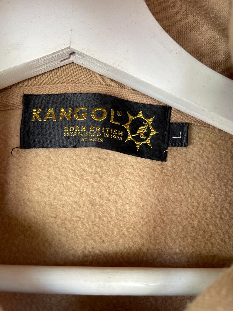 May include: Close-up of a tan-colored hoodie with a black label. The label reads "KANGOL" in gold lettering, with "BORN BRITISH ESTABLISHED IN 1938" below. A kangaroo logo is on the right side of the label. The size is "L".