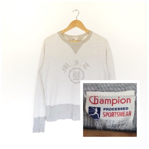 Puede incluir: Una sudadera Champion gris de manga larga con un diseño de bloques de color gris y blanco. La sudadera tiene cuello redondo y presenta un logotipo bordado de estilo vintage que dice "M.I.T." con un escudo. La etiqueta del logotipo de Champion es visible en la esquina inferior derecha de la sudadera.