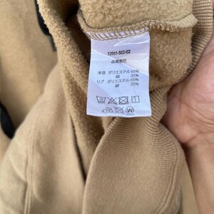 May include: Close-up of a tan sweatshirt with a tag. The tag displays the number 12931-502-02 and Japanese text indicating the material composition: 65% polyester and 35% cotton. Washing instructions are also visible.