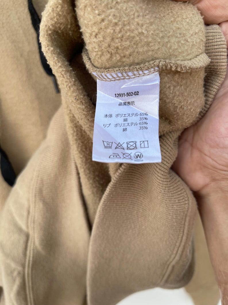 May include: Close-up of a tan sweatshirt with a tag. The tag displays the number 12931-502-02 and Japanese text indicating the material composition: 65% polyester and 35% cotton. Washing instructions are also visible.