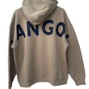 May include: Tan hoodie with the word "ANGO." printed in large, black letters across the back. The hoodie has a hood and long sleeves. The garment is hanging from a metal hanger.