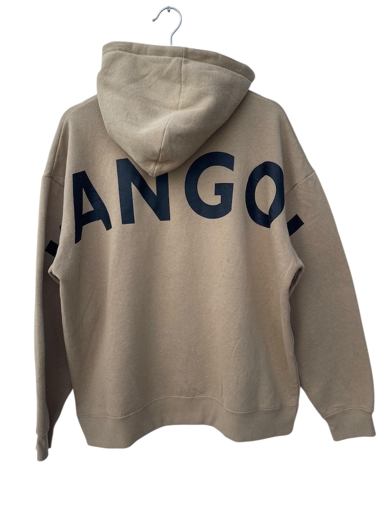 May include: Tan hoodie with the word "ANGO." printed in large, black letters across the back. The hoodie has a hood and long sleeves. The garment is hanging from a metal hanger.