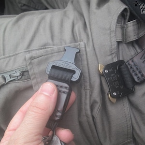 May include: A black and silver belt buckle with the text "Wolf Wind Leatherworks" and a paw print. The buckle is attached to a black strap with a black and silver clip.