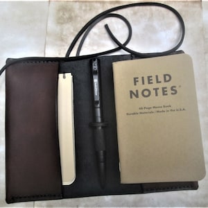 Może przedstawiać: Brązowy skórzany organizer na notesy z czarnym długopisem, małym blokiem notesowym i notesem Field Notes. Notes jest brązowy z tekstem "FIELD NOTES" i "48-Page Memo Book Durable Materials/Made in the U.S.A."