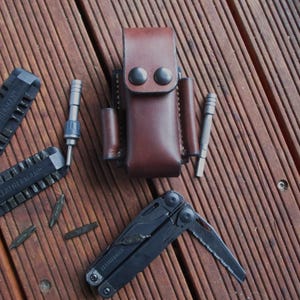 May include: A brown leather multi-tool case with a black multi-tool, screwdriver bits, and a screwdriver. The tools are arranged on a wooden surface. The multi-tool has a saw blade.