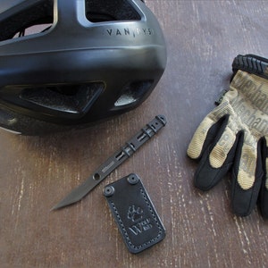 Puede incluir: Un casco de bicicleta negro con la palabra "VAN DYS" en el lateral, un cuchillo de uso general negro con una funda de cuero negro y un par de guantes de trabajo de color beige y negro.