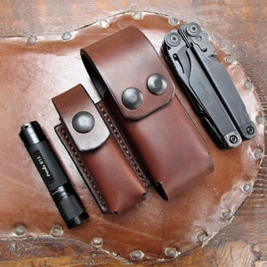 May include: Two brown leather holsters with snap closures, one for a flashlight and one for a multi-tool. The flashlight is black and has the text "Peak 112" on the side. The multi-tool is black and has the text "Leatherman" on the side.