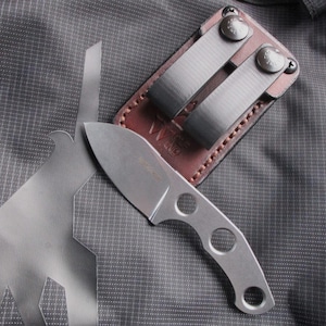 Op de afbeelding: Een klein, zilverkleurig mes met drie cirkelvormige uitsparingen in het handvat ligt op een gestructureerd grijs oppervlak. Het mes wordt gecombineerd met een bruine leren schede met twee metalen clips. De afbeelding is een close-up.