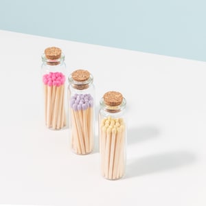 May include: Three glass jars filled with colorful matches. The jars have cork lids and are arranged in a row on a white surface. The matches are pink, purple, and yellow.