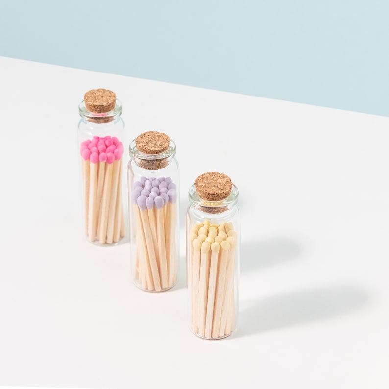 May include: Three glass jars filled with colorful matches. The jars have cork lids and are arranged in a row on a white surface. The matches are pink, purple, and yellow.