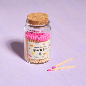 May include: A clear glass jar filled with pink-tipped matches, topped with a cork lid. The jar has a label that reads "enlighten the occasion spark joy" with star and dot graphics. Two matches lie beside the jar on a purple surface.