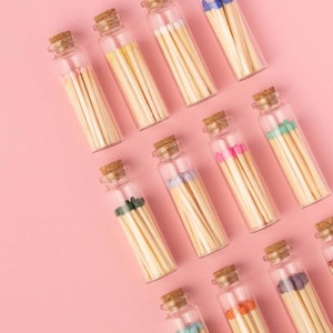May include: A collection of glass jars filled with colorful matches on a pink background. The jars are arranged in a staggered pattern, with the tops of the jars facing the viewer. The matches are all different colors, including blue, green, pink, and orange.