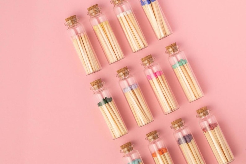 May include: A collection of glass jars filled with colorful matches on a pink background. The jars are arranged in a staggered pattern, with the tops of the jars facing the viewer. The matches are all different colors, including blue, green, pink, and orange.