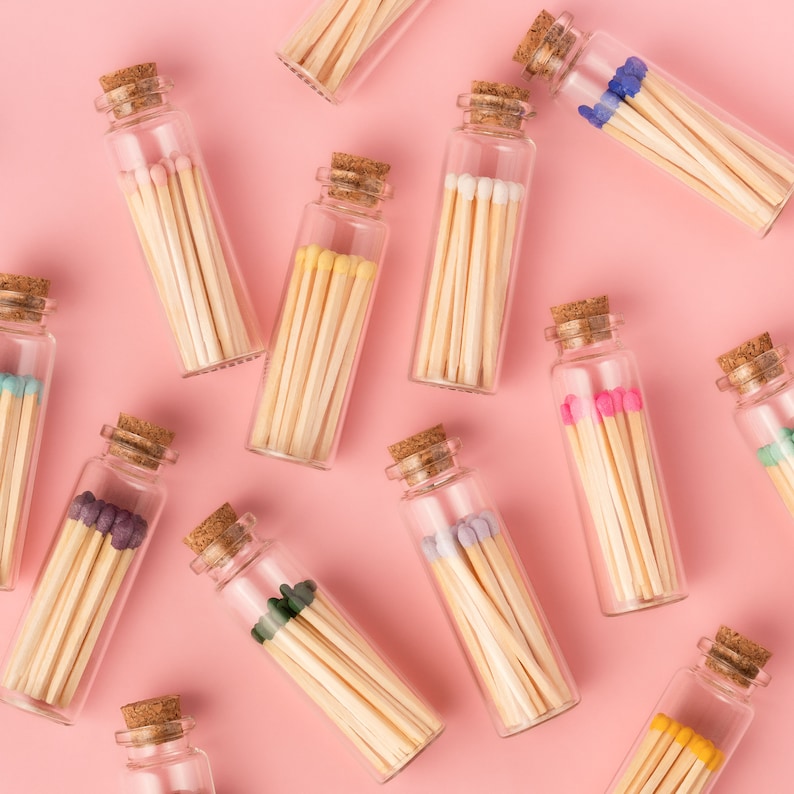 May include: A collection of glass bottles filled with colorful matchsticks. The bottles have cork tops and are arranged on a pink background.