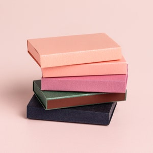 May include: Stack of four boxes in different colors, including pink, peach, purple, green, and black. The boxes are all rectangular and have a smooth, textured surface.