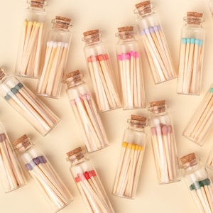 May include: Assortment of small glass bottles filled with colorful matchsticks. The bottles have cork tops and are arranged on a light beige background.